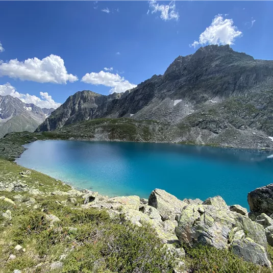 Ein klarer, turquoise See umgeben von hohen Bergen und grünem Gras. Der Himmel ist blau mit einigen weißen Wolken.