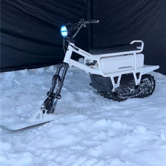 Ein innovatives Schnee-Motorrad steht auf einem schneebedeckten Boden. Die Umgebung ist von Dunkelheit umgeben, während das Fahrzeug eine helle Farbe hat.
