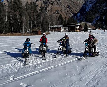 Eine Gruppe von vier Personen fährt auf speziellen Schneefahrzeugen im Schnee. Im Hintergrund sind Berge und ein Gebäude sichtbar.