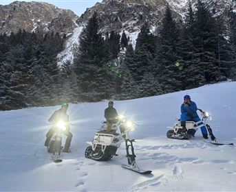 Three people are riding on special snowbikes in the snow. In the background, snow-covered mountains and pine trees can be seen.
