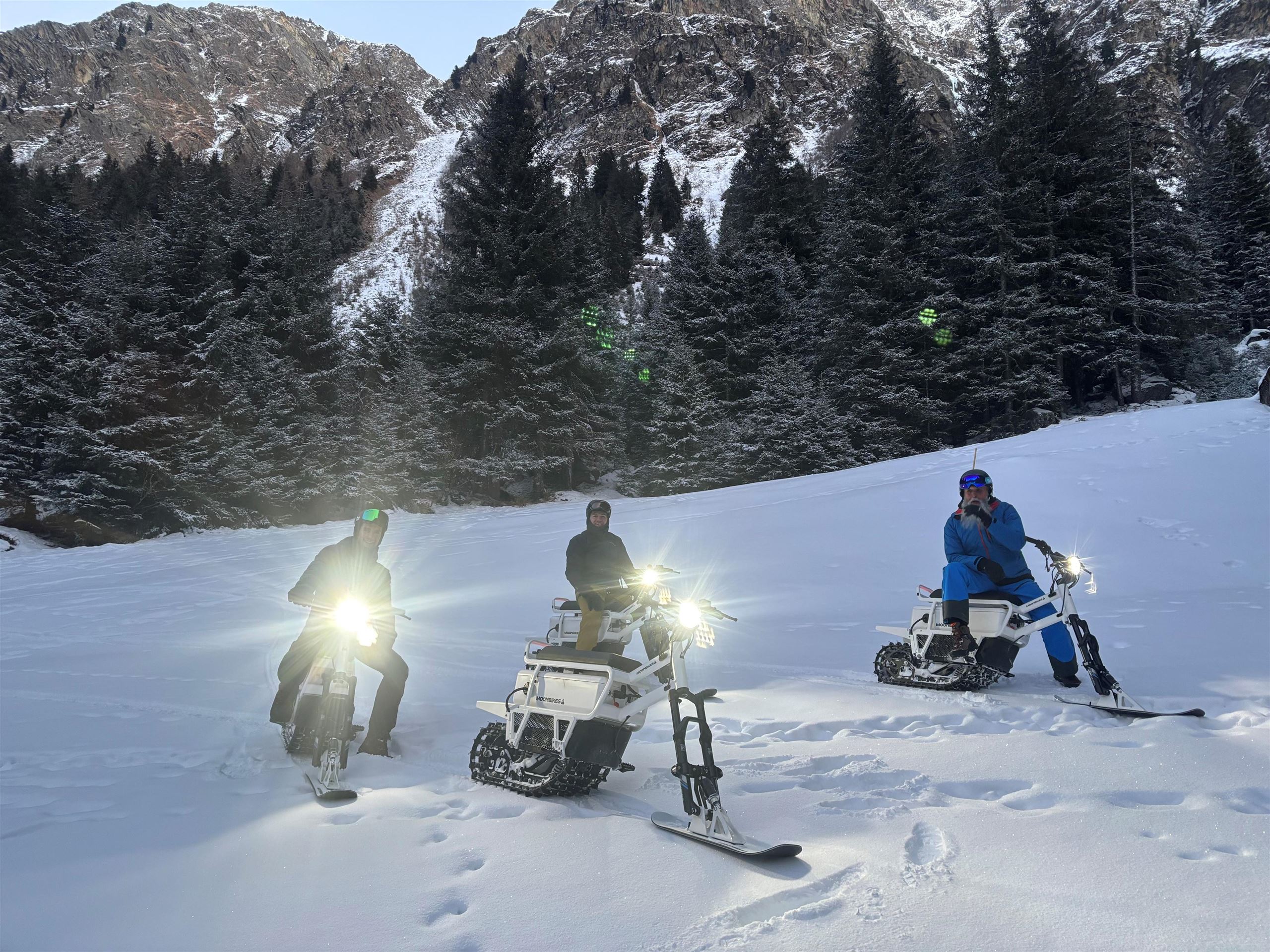 Drei Personen fahren auf speziellen Snowbikes im Schnee. Im Hintergrund sind schneebedeckte Berge und Tannenbäume zu sehen.