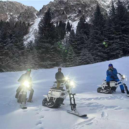 Drei Personen fahren auf speziellen Snowbikes im Schnee. Im Hintergrund sind schneebedeckte Berge und Tannenbäume zu sehen.