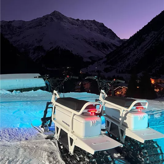 Zwei Schneemobile stehen auf einem schneebedeckten Hang bei Dämmerung. Im Hintergrund sind beeindruckende Berge und ein klarer Himmel sichtbar.