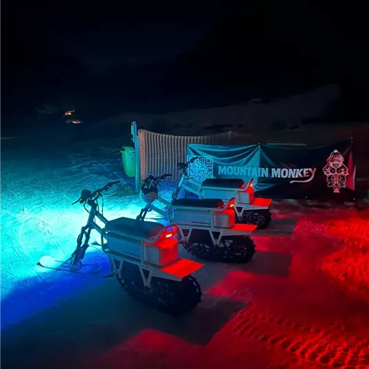 Drei E-Bikes stehen bei Nacht auf einem beleuchteten Gelände. Im Hintergrund ist ein Banner mit der Aufschrift "MOUNTAIN MONKEY" zu sehen.