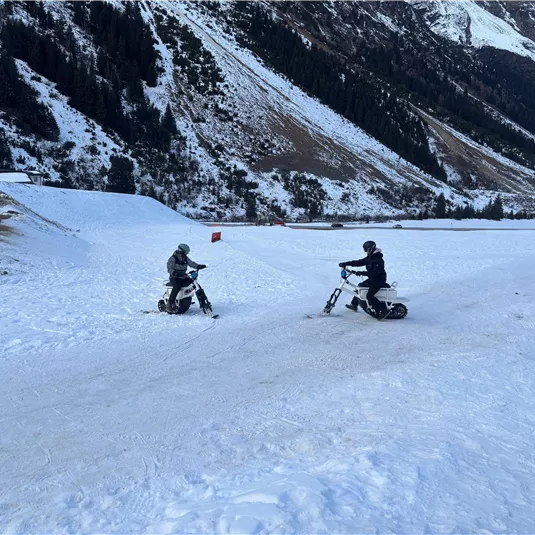 Zwei Personen fahren auf Schneescootern in einer winterlichen Berglandschaft. Im Hintergrund sind schneebedeckte Berge und eine ruhige, verschneite Umgebung zu sehen.