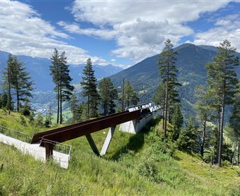 Eine moderne Aussichtsplattform in den Bergen, umgeben von Bäumen. Im Hintergrund sind majestätische Berge und ein blauer Himmel zu sehen.