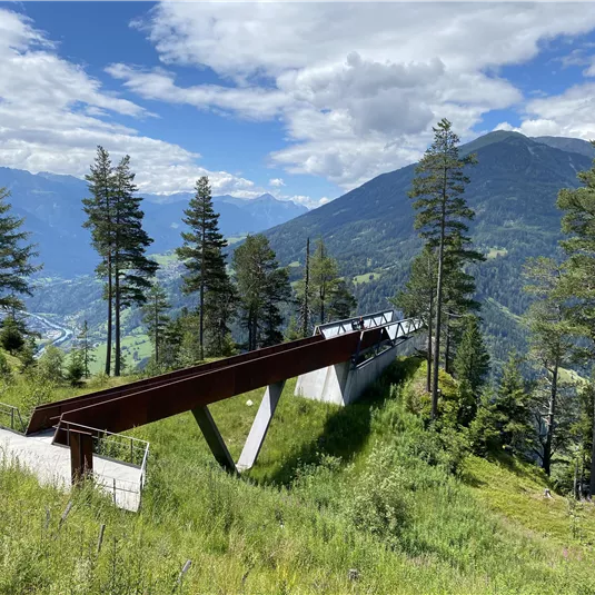 Eine moderne Aussichtsplattform in den Bergen, umgeben von Bäumen. Im Hintergrund sind majestätische Berge und ein blauer Himmel zu sehen.