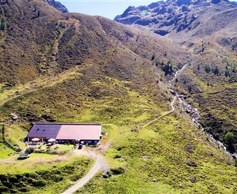 A picturesque mountain cabin surrounded by green forests and majestic mountains. The sky is blue with a few white clouds.