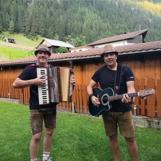 Zwei Musiker stehen im Garten, einer spielt Akkordeon und der andere Gitarre. Im Hintergrund sind Holzschuppen und grüne Wiesen zu sehen.