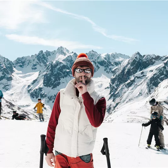A wintry ski landscape with snow-covered mountains. A person in warm clothing is making a peace sign, while other skiers can be seen in the background.