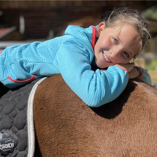 A smiling girl is lying relaxed on the back of a horse. In the background, wooden houses and a green environment are visible.