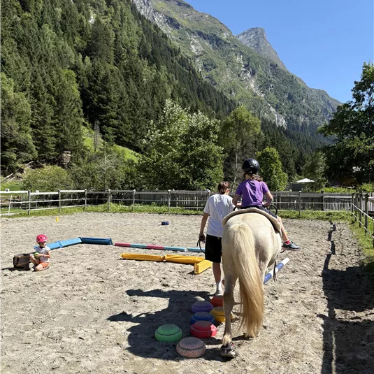 A riding arena amidst mountains, where a child is sitting on a horse. In the background, trees and a clear blue sky are visible.
