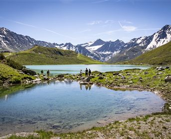 A picturesque mountain lake with sparkling water and snow-covered peaks in the background. Green grass and rocks surround the shore, while people are standing by the water.