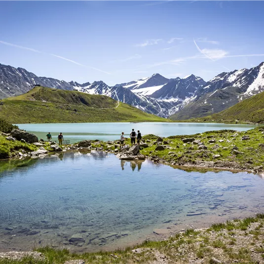 Ein malerischer Bergsee mit glitzertem Wasser und schneebedeckten Gipfeln im Hintergrund. Grünes Gras und Felsen umgeben das Ufer, während Menschen am Wasser stehen.
