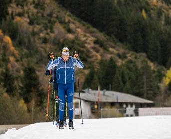 Eine Gruppe von Skiläufern bewegt sich auf einer verschneiten Strecke. Im Hintergrund sind Bäume und ein Gebäude zu sehen.
