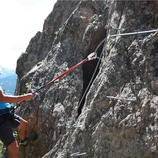 A man is climbing a steep rock face with safety hooks. In the background, green forests and mountains can be seen.