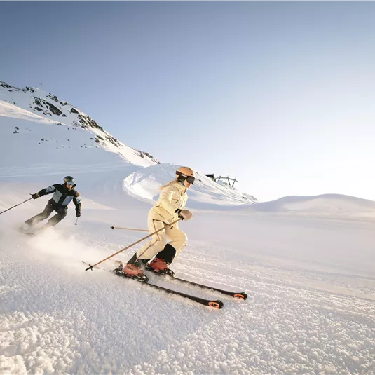 Two skiers are skiing down a snowy slope. The sky is clear and the sun is shining.