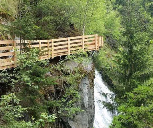 A wooden path runs along a steep slope with lush greenery. A waterfall flows down beside the path.