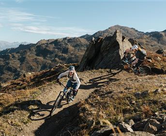 Eine Person steht auf einem Hügel und blickt auf die beeindruckende Berglandschaft. Im Hintergrund ist ein See und ein klarer Himmel mit einigen Wolken zu sehen.