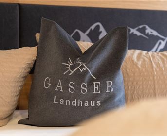 Ein dekoratives Kissen mit der Aufschrift "GASSER Landhaus". Es liegt auf einem Bett in einem gemütlichen Raum.