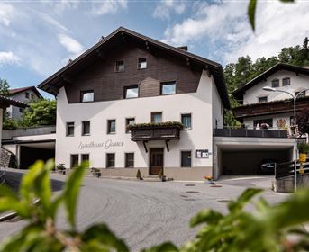 Ein charmantes Gebäude mit einem rustikalen Dach und hellen Wänden. Es liegt an einer ruhigen Straßenkreuzung, umgeben von grünen Bäumen und Bergen.