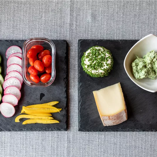 A healthy snack arrangement with fresh vegetable strips, cherry tomatoes, and various dips. On the right side, there are slices of cheese and a herb quark.