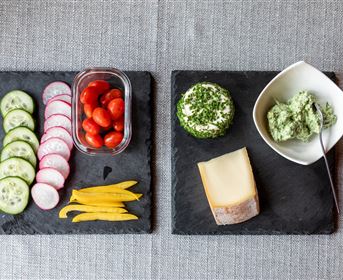 A healthy snack arrangement with fresh vegetable strips, cherry tomatoes, and various dips. On the right side, there are slices of cheese and a herb quark.