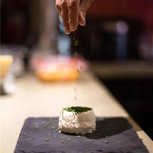 A hand sprinkles herbs over a small cream cheese cookie on a black plate. The background shows a blurry kitchen environment.