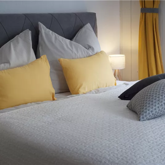 Ein modernes Schlafzimmer mit einem Bett, das mit bunten Kissen dekoriert ist. Sanftes Licht und helle Farben schaffen eine einladende Atmosphäre.