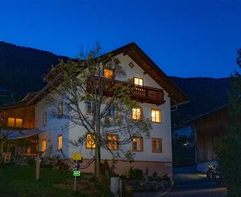 Ein gemütliches Haus mit beleuchteten Fenstern, umgeben von Bäumen und Bergen. Die Abenddämmerung schafft eine einladende Atmosphäre.