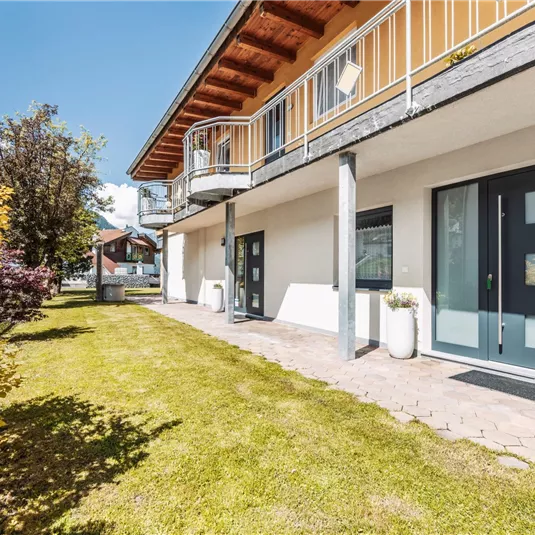 A modern building with a spacious terrace and large windows. The well-maintained garden in the foreground is adorned with colorful shrubs.