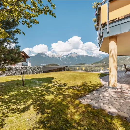 A peaceful landscape with a well-kept garden and a view of the mountains. The sky is blue with some white clouds.