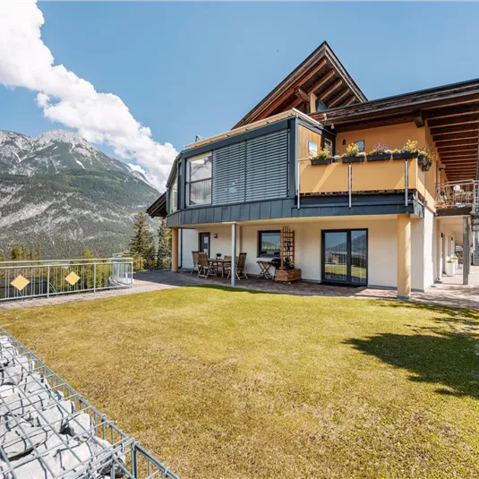A modern house with large windows and a terrace. In the background, impressive mountains and a blue sky can be seen.