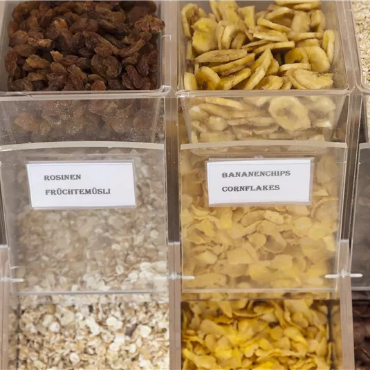Various types of crunchy muesli and cereals in transparent containers. The ingredients are clearly labeled and range from nuts to banana chips.