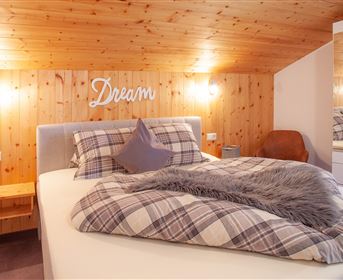 A cozy bedroom with a large bed and wooden paneling. On the bed lies a soft blanket and a pillow.