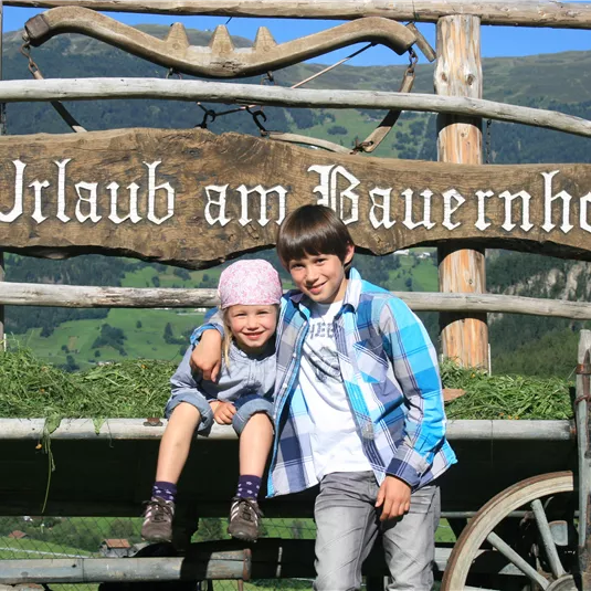 Zwei Kinder lächeln vor einem Holzschild mit der Aufschrift "Urlaub am Bauernhof". Im Hintergrund sind grüne Wiesen und Berge zu sehen.