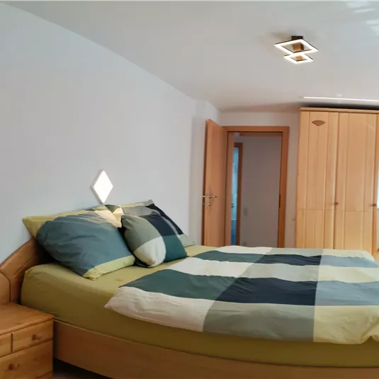 A spacious bedroom with a large bed and a simple desk. The walls are light and there is a large wooden wardrobe.