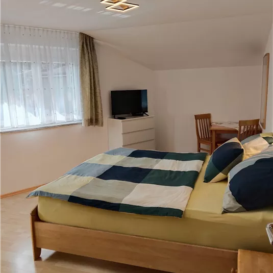 A cozy bedroom with a large bed and colorful blankets. In the background, there is a television and a small dining area.