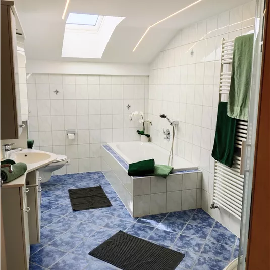 A bright bathroom with a bathtub and a window. The walls are tiled white, and there are green towels and bath mats.