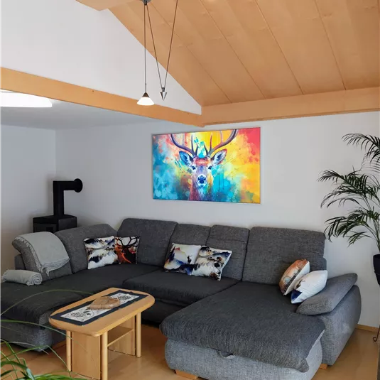 A cozy living room with a large gray couch and a colorful mural of a deer. Next to the couch are a small table and some plants.