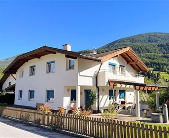 Ein modernes Haus mit einem versetzten Dach und blauen Fensterläden. Im Hintergrund sind grüne Berge und ein klarer blauer Himmel zu sehen.