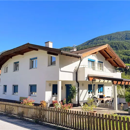 A modern house with a staggered roof and blue shutters. In the background, green mountains and a clear blue sky can be seen.