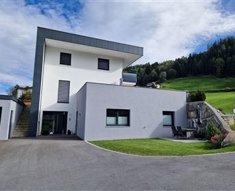 Ein modernes Haus mit zwei Etagen und einem klaren, minimalistischen Design. Im Hintergrund sind grüne Hügel und ein blauer Himmel zu sehen.