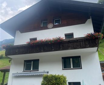 Ein traditionelles alpines Haus mit einem Holzbalkon und bunten Blumen. Der Himmel ist bewölkt und es ist eine grüne Landschaft im Hintergrund sichtbar.