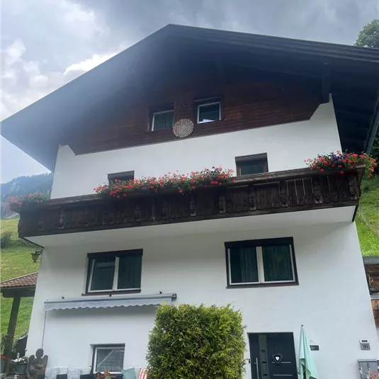 A traditional alpine house with a wooden balcony and colorful flowers. The sky is cloudy and a green landscape is visible in the background.