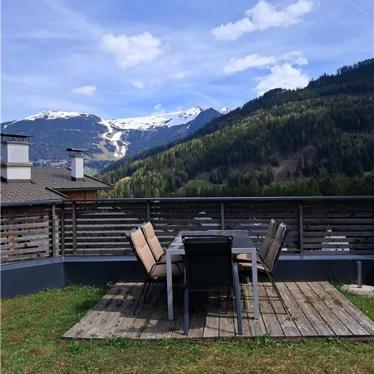 A beautiful terrace with a large table and chairs. In the background are picturesque mountains with snow-covered peaks.