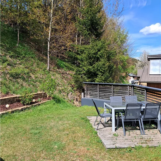 A well-maintained garden with a wooden seating area and a table with chairs. In the background, trees and a blue sky are visible.