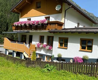 Ein malerisches Haus in den Bergen mit einem Balkon voller Blumen. Der Garten ist grün und einladend, umgeben von der schönen Natur.