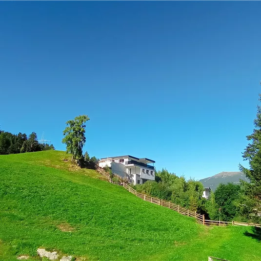 A green hill with a white house and trees. The sky is clear and blue.