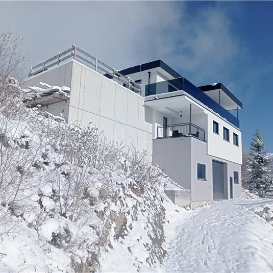 A modern house on a snow-covered hill. The sky is clear and blue, surrounded by a wintry landscape.
